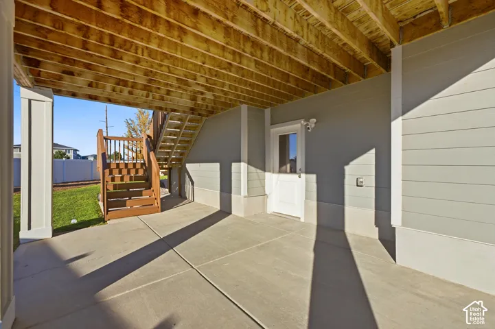 Covered concrete patio with exposed wood joist ceiling, featuring wood-finish stairs, and a white exterior door