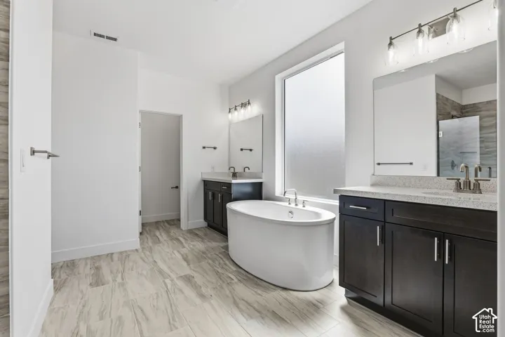 Freestanding soaking tub, dual vanity with dark wood cabinetry, light-toned stone countertops, large frosted window, and wood-finish tile flooring