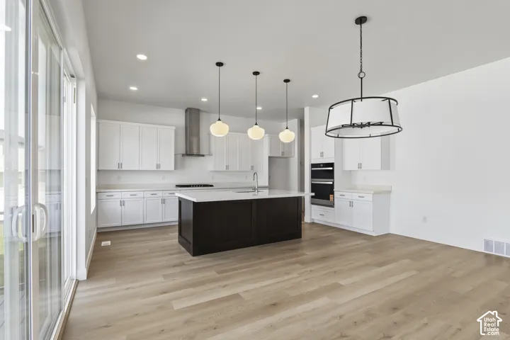Modern kitchen with a dark-toned island, light wood-finish flooring, white cabinetry, integrated double ovens, and a stainless steel range hood