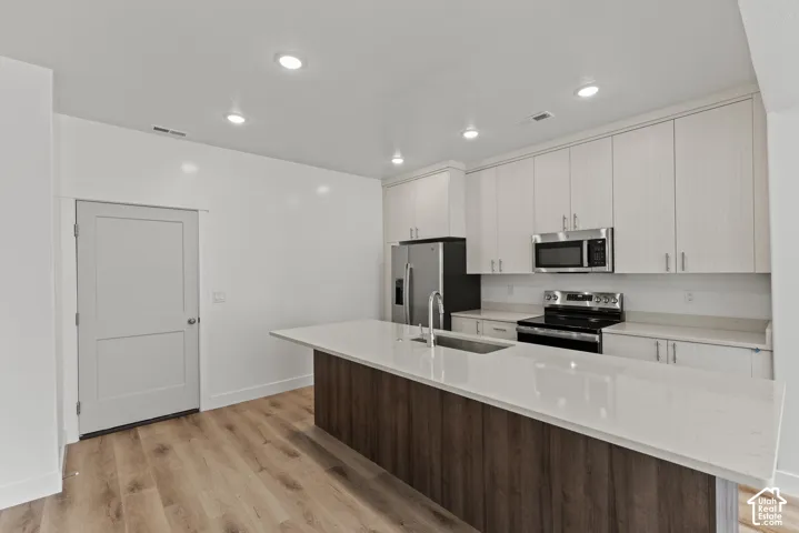 Kitchen featuring wood-finish flooring, recessed lighting, white perimeter cabinetry, stainless steel appliances, and a central island with a sink and white countertop