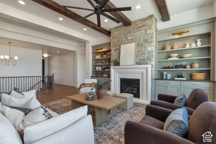 Stone-clad fireplace with a classic mantel, built-in shelving, wood-finish flooring, exposed ceiling beams, and a ceiling fan with integrated lighting