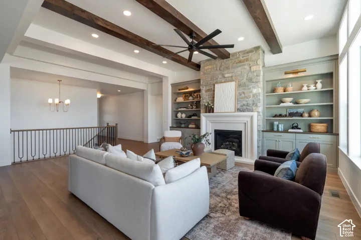 Exposed wood beams accentuating the ceiling, stone fireplace with a mantel, built-in shelving with lower cabinetry, wood-finish flooring, and large windows