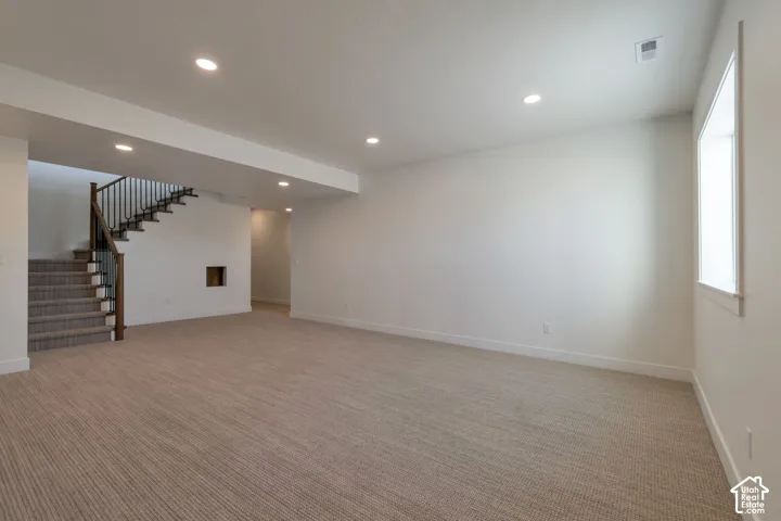 Expansive carpeted area with recessed lighting, a staircase featuring dark wood and metal balusters, and a window providing natural illumination