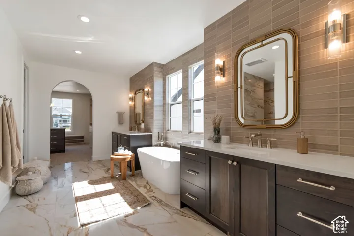 Freestanding tub with a wall-mounted faucet, dual vanities featuring dark wood cabinetry and white countertops, and a tiled accent wall
