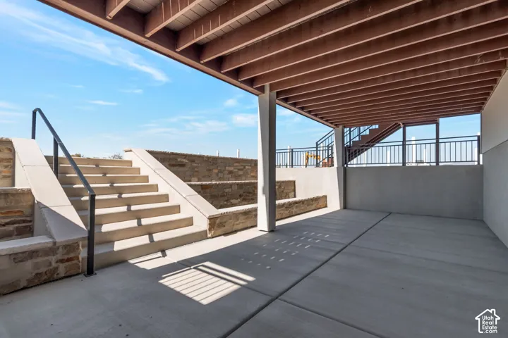Covered concrete patio with exposed beam ceiling, concrete stairs with black metal handrails, and a stone-clad retaining wall