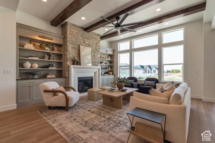 Living area featuring a stone-clad fireplace with a white mantel, built-in shelving, wood-finish flooring, exposed ceiling beams, and large picture windows