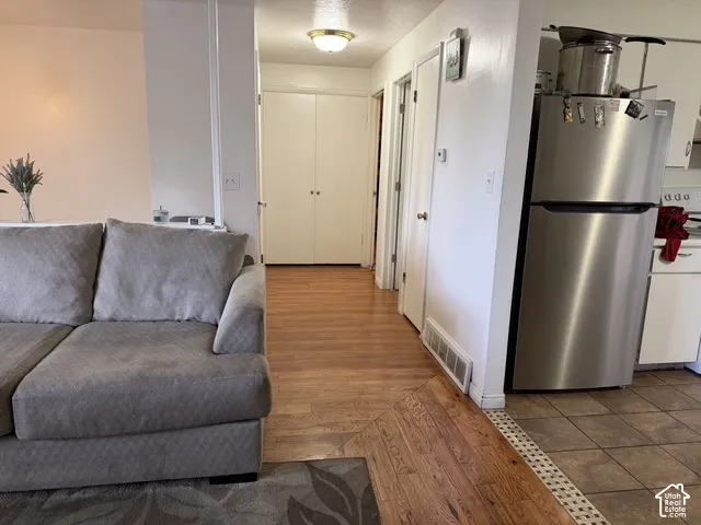 Warm wood-finish flooring extending into the hallway, complemented by a kitchen area with tile flooring and a stainless steel refrigerator