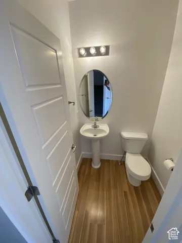 Pedestal sink, oval mirror, wall-mounted light fixture, wood-finish flooring, and a white toilet
