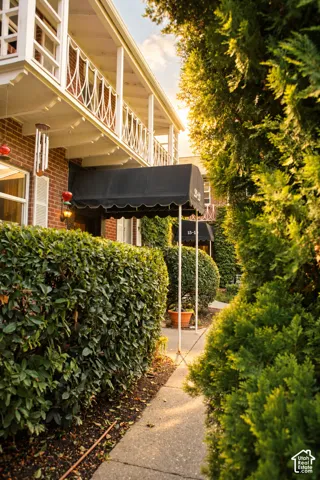 Brick exterior with white balcony railings, black fabric awning, and a concrete pathway surrounded by manicured hedges
