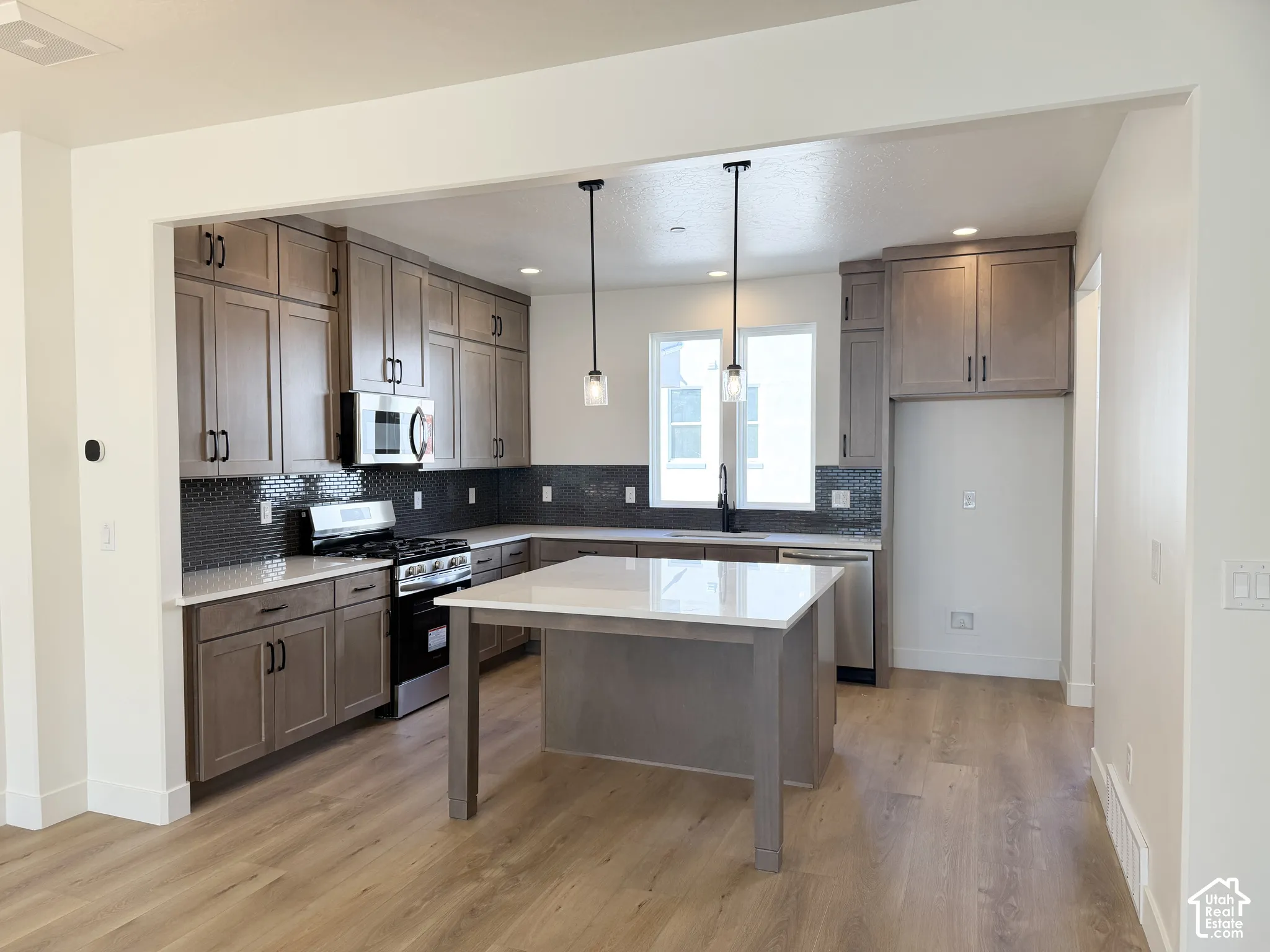 Kitchen featuring stainless steel appliances, wood-finish flooring, dark tile backsplash, light wood cabinetry, and a central island with a white countertop