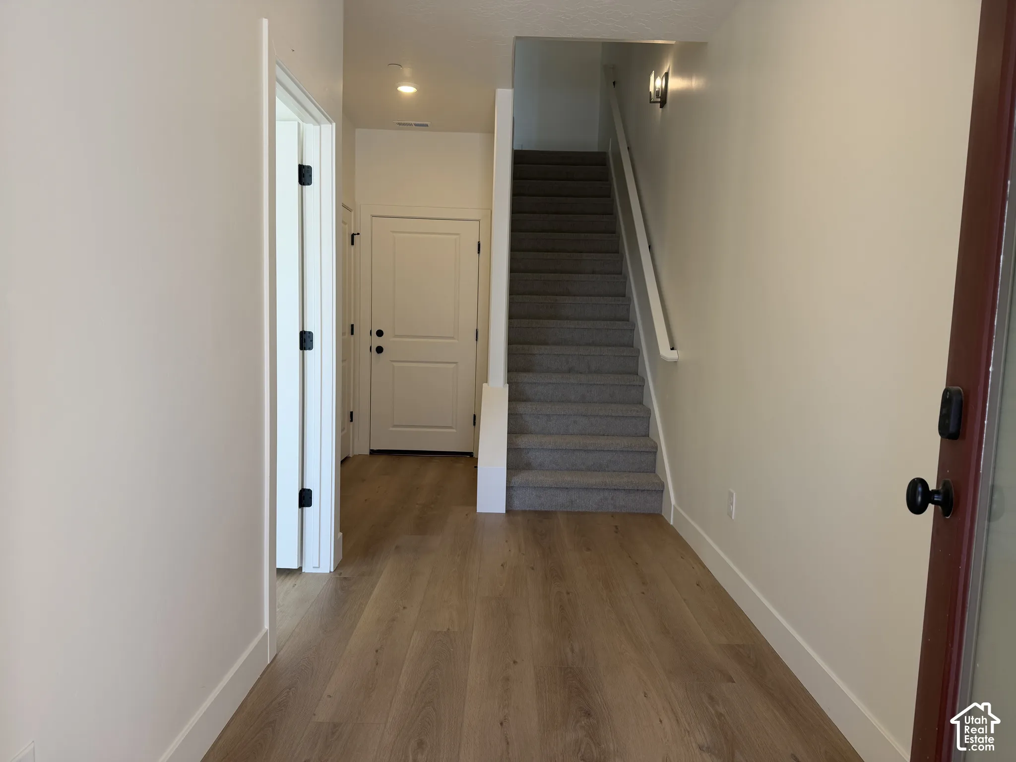Wood-finish flooring, staircase with carpeted treads, wall-mounted light fixture, recessed ceiling lighting, and solid interior doors with dark hardware