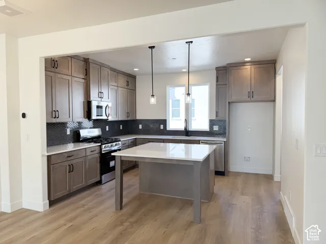 Kitchen featuring stainless steel appliances, wood-finish flooring, dark tile backsplash, light wood cabinetry, and a central island with a white countertop