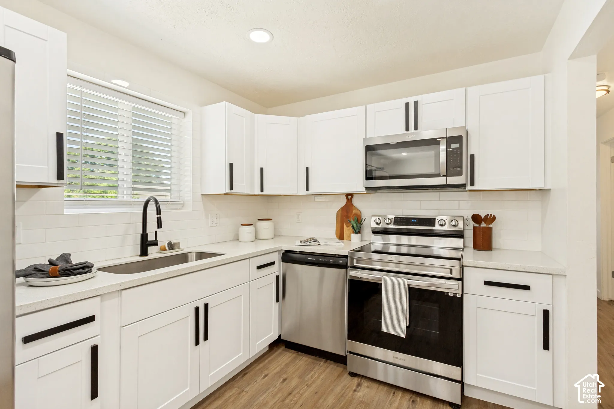 White shaker-style cabinetry with matte black hardware, stainless steel appliances, durable wood-finish flooring, white subway tile backsplash, and recessed lighting