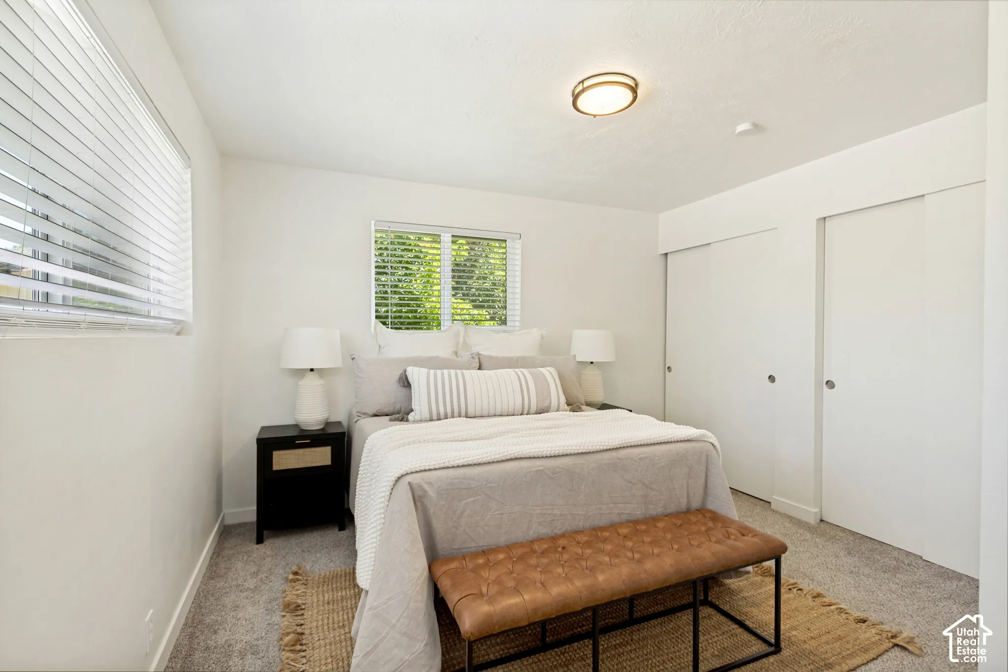 Plush carpet flooring, bright white walls, two windows with horizontal blinds, flush mount ceiling light, and dual sliding closet doors