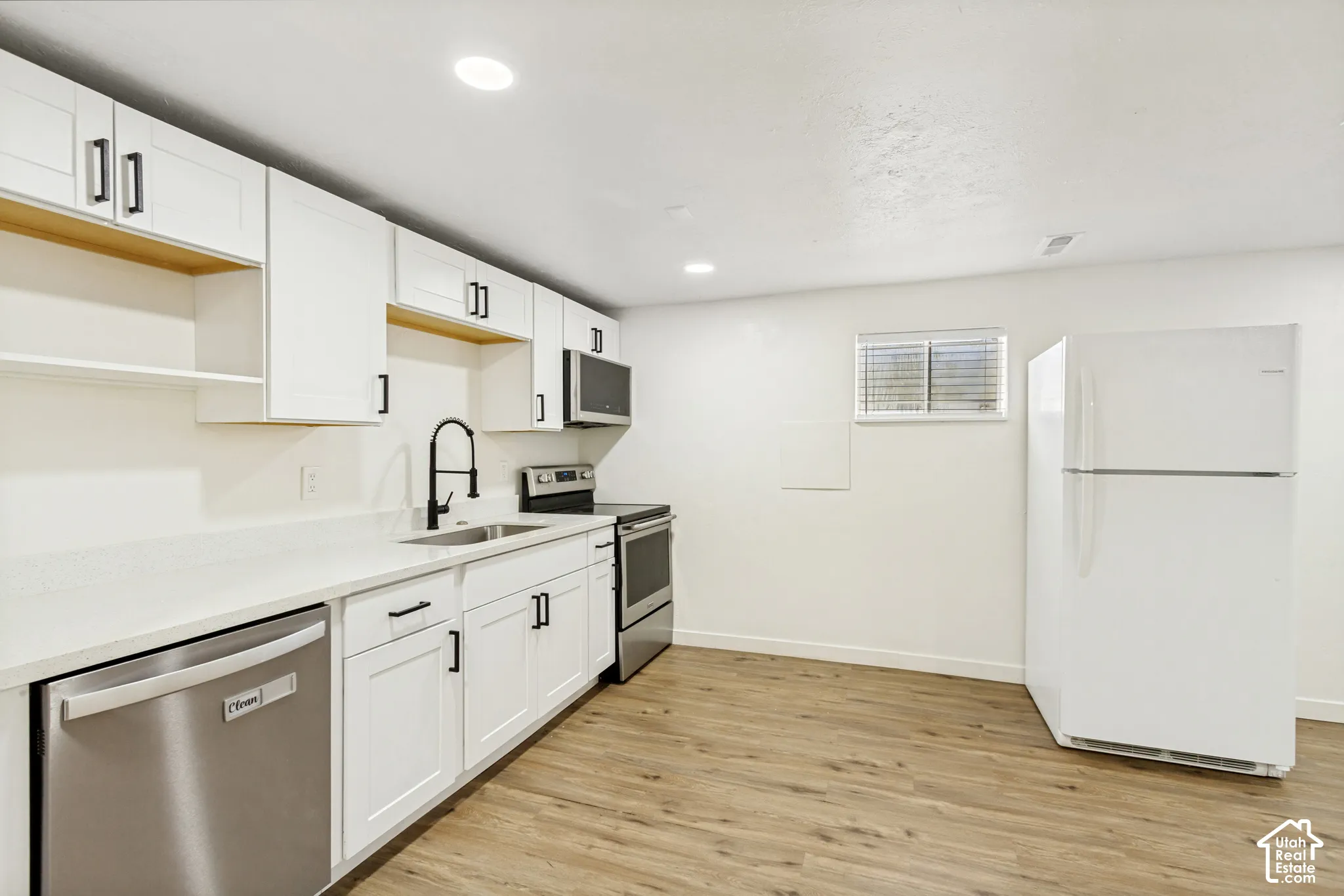 White cabinetry with black hardware, stainless steel appliances, wood-finish flooring, and a window with blinds