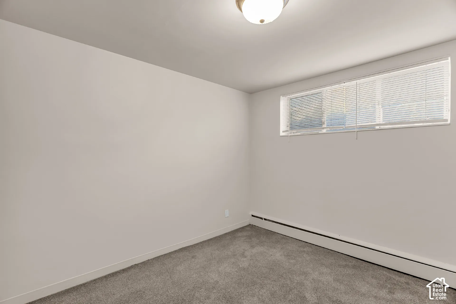 Light gray wall color, low-profile ceiling light fixture, horizontal window with blinds, baseboard heating unit, and gray carpet