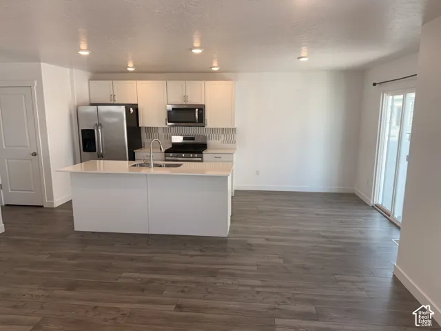 Open-concept kitchen featuring an island with a built-in sink, stainless steel appliances, white cabinetry, and wood-finish flooring