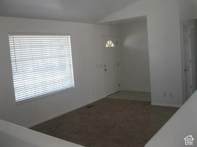 Vaulted ceiling, neutral wall paint, carpet flooring, window with blinds, and white entry door with decorative glass