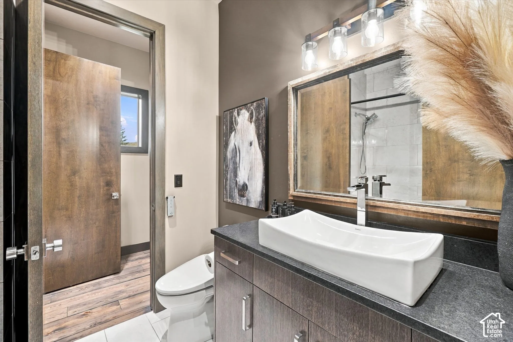 Wood-finish bathroom door, white rectangular vessel sink, dark wood-finish vanity with drawer storage, sleek chrome faucet, and contemporary vanity lighting