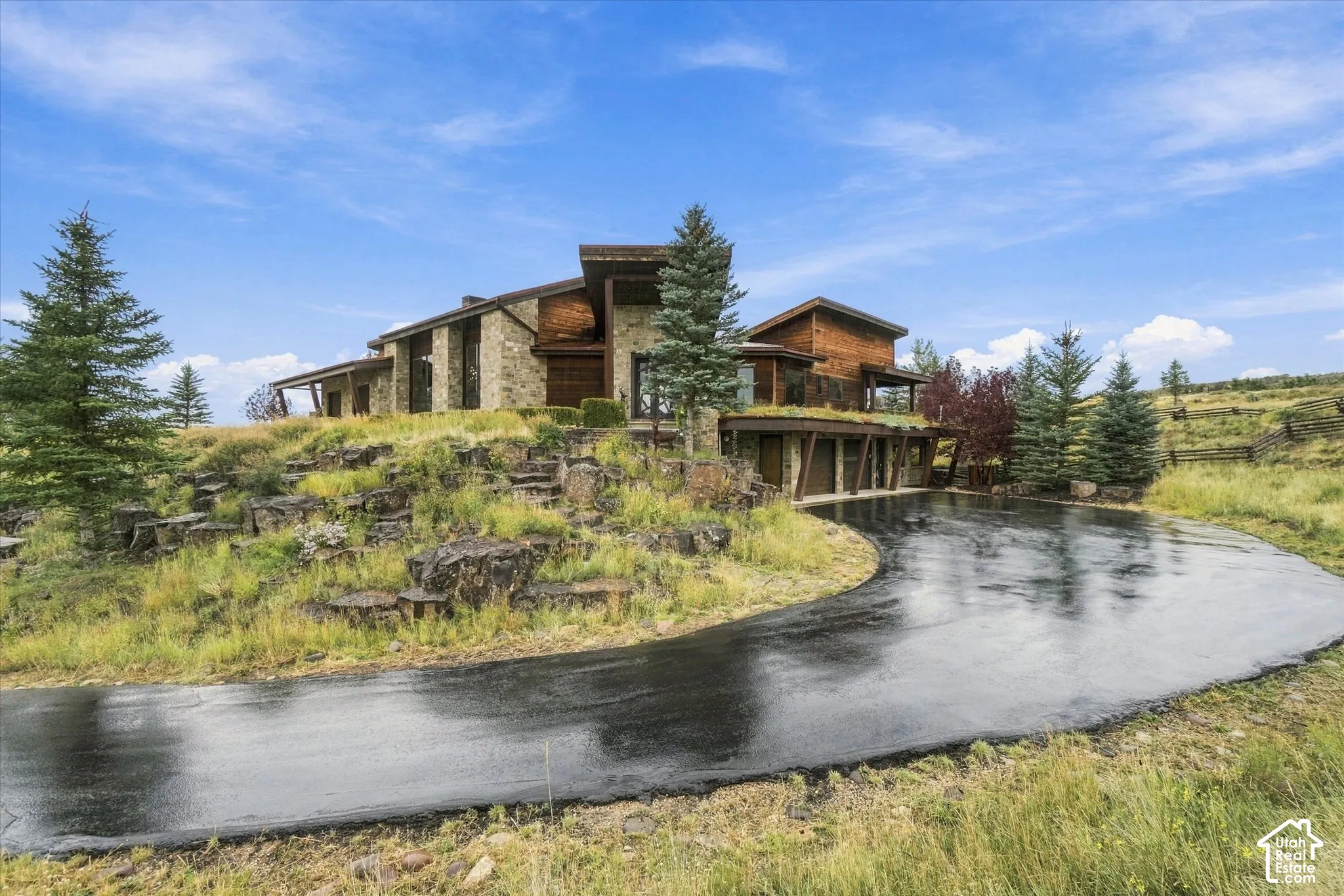 Stone and wood-clad exterior with multiple architectural levels, natural landscaping, an expansive paved driveway, and mature evergreen trees