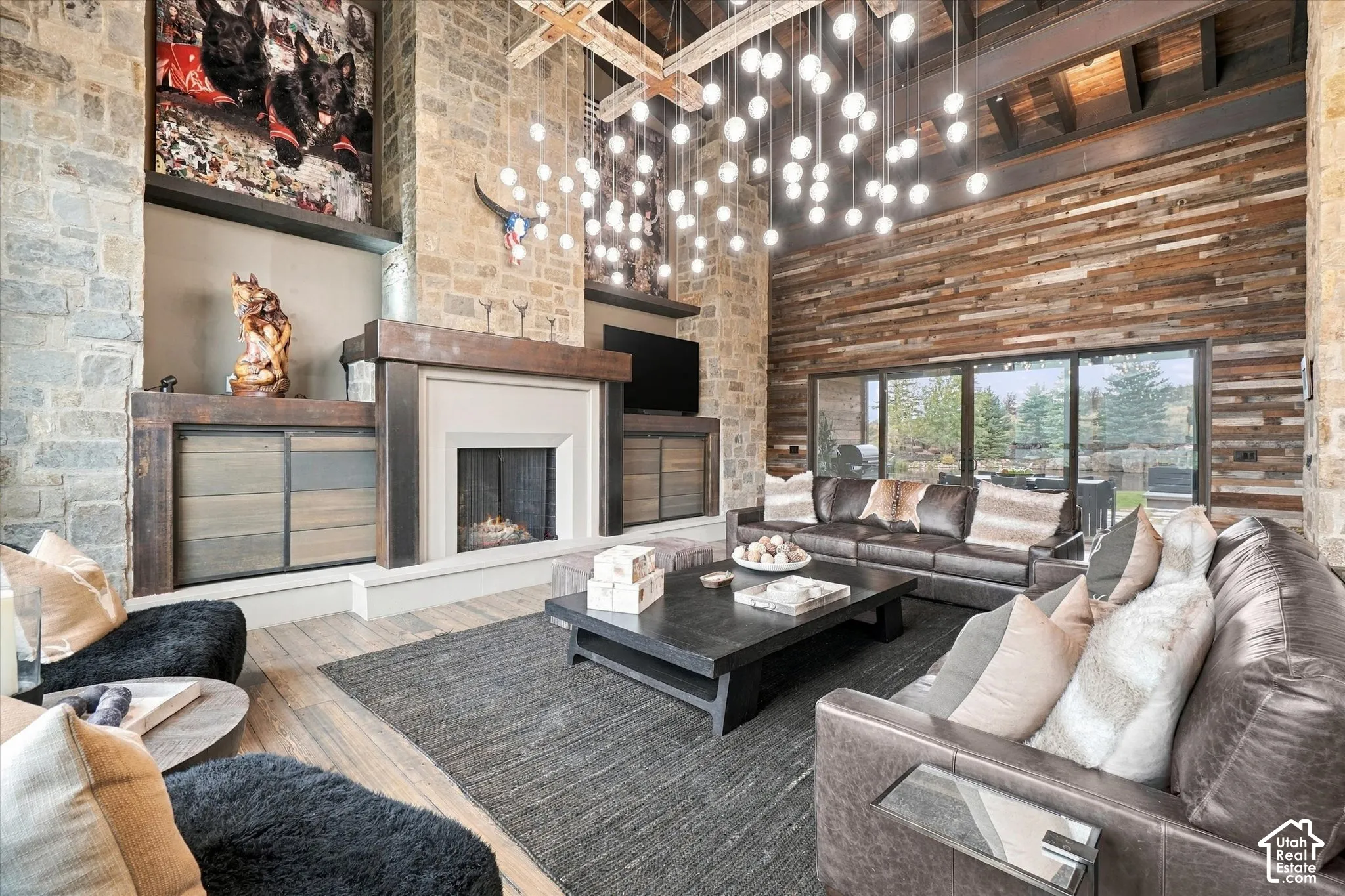 Vaulted ceiling with exposed wood beams, stone-clad fireplace with mantel, wood-finish flooring, full-height stacked wood accent wall, and sliding glass doors