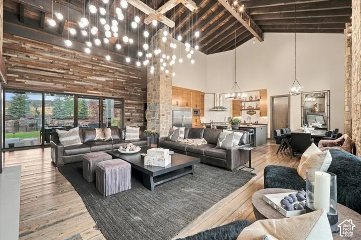 Vaulted ceiling with exposed wood beams, expansive wood-finish flooring, full-height stone wall, large sliding glass doors, and recessed kitchen with stainless steel appliances