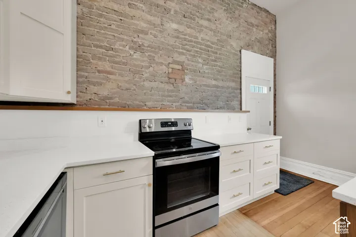 Exposed brick wall, stainless steel range, white cabinetry with gold-tone hardware, white countertops, and wood-finish flooring