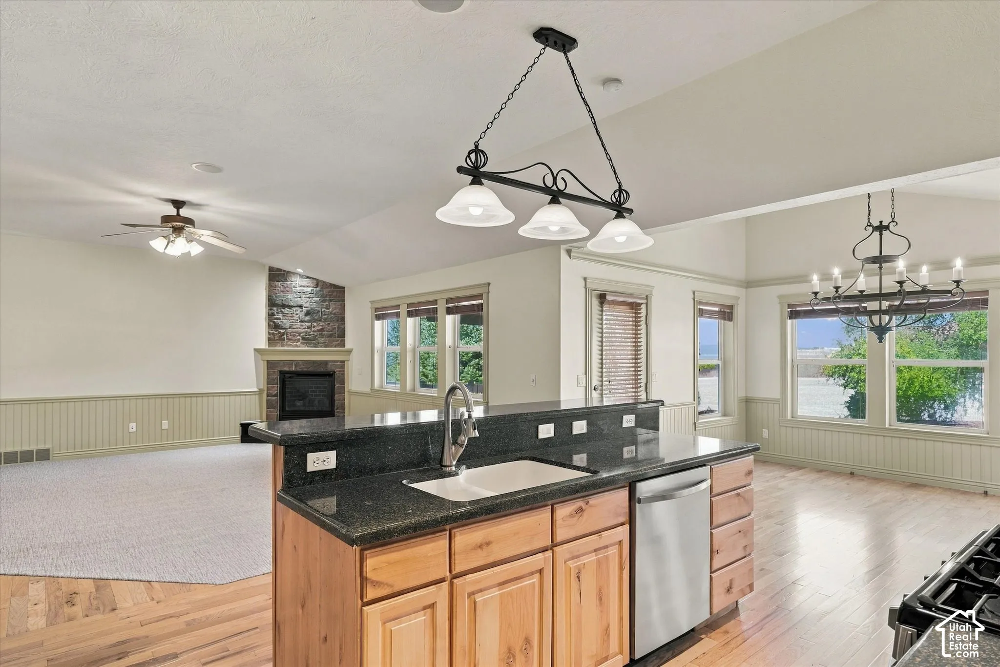 Kitchen island with dark countertop and undermount sink, stainless steel dishwasher, vaulted ceilings, stone-faced fireplace, and wood-finish flooring