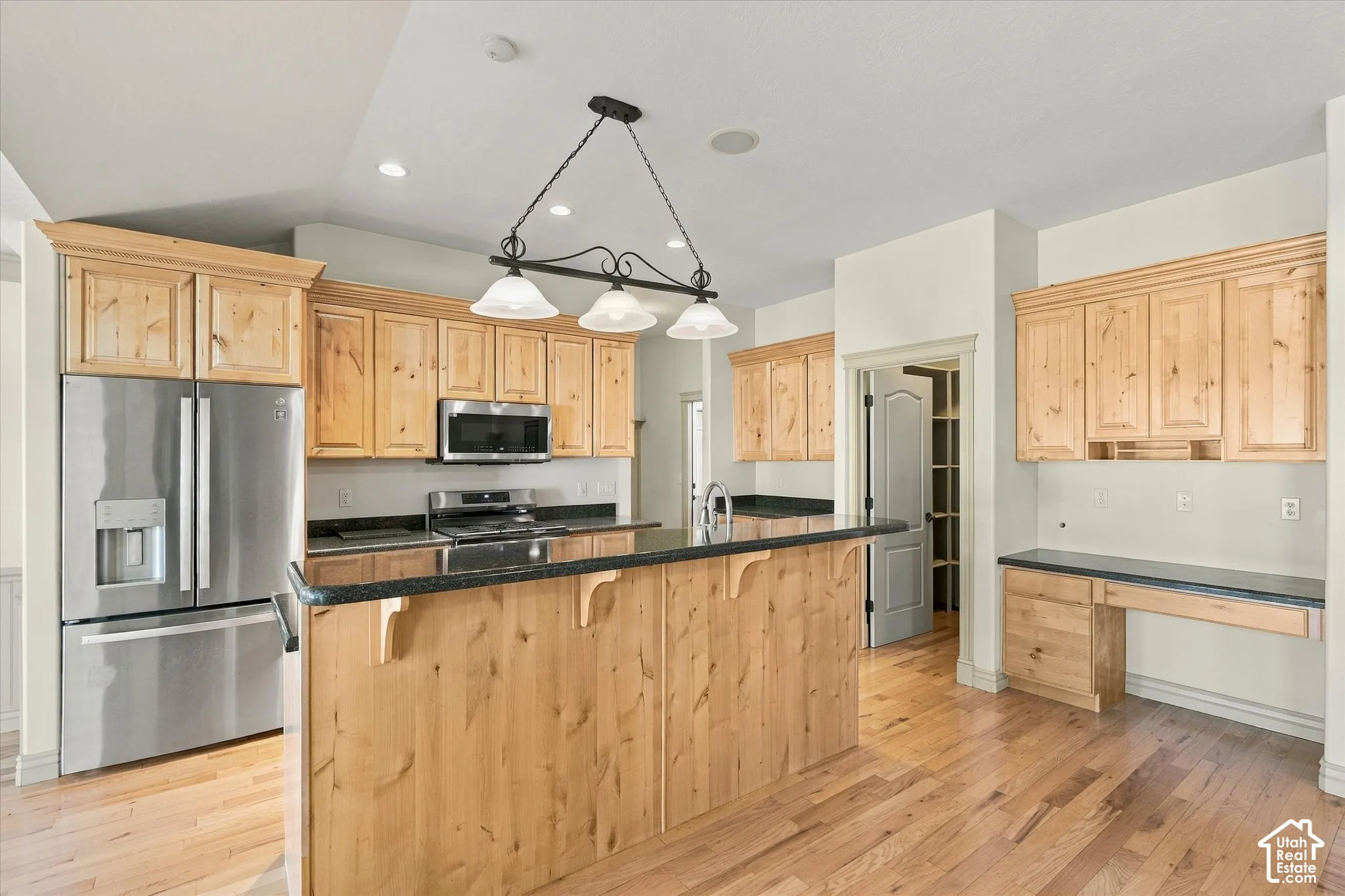 Kitchen featuring hardwood flooring, natural wood cabinetry, stainless steel appliances, dark countertops, and a built-in desk area