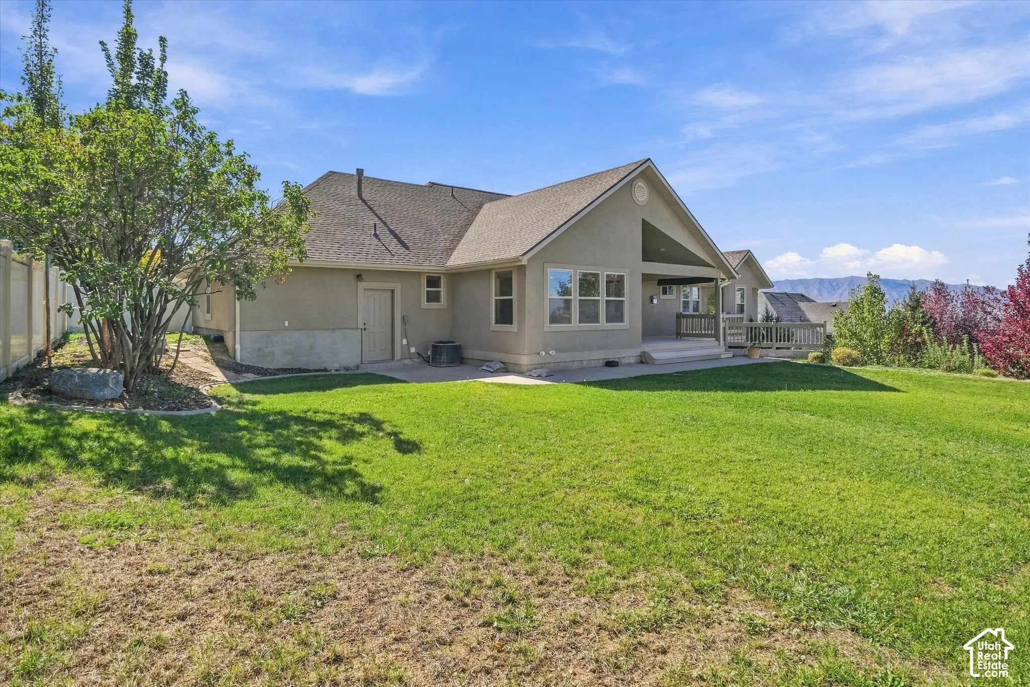 Expansive green lawn, stucco exterior, covered patio, rear decking, and sloped landscaping with mature trees