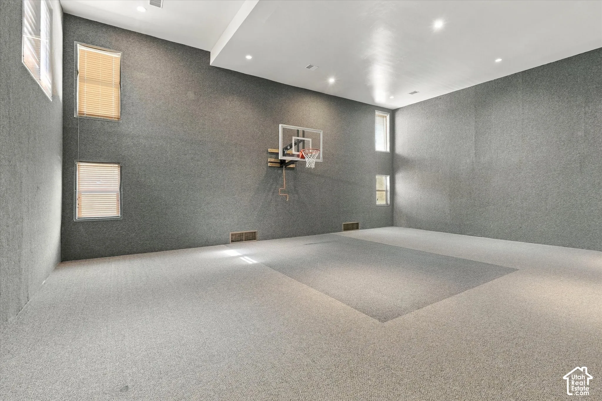 Expansive room featuring recessed lighting, a basketball hoop, and multiple windows with blinds, all set against textured wall coverings and light-toned carpeting with a distinct court outline