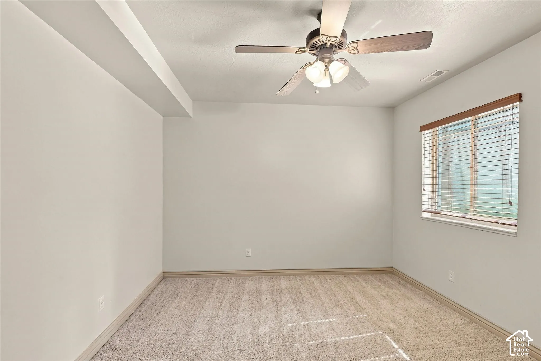 Recessed ceiling beam with flush lighting, ceiling fan with integrated lighting, window with wood-finish blinds, and neutral-toned carpet flooring