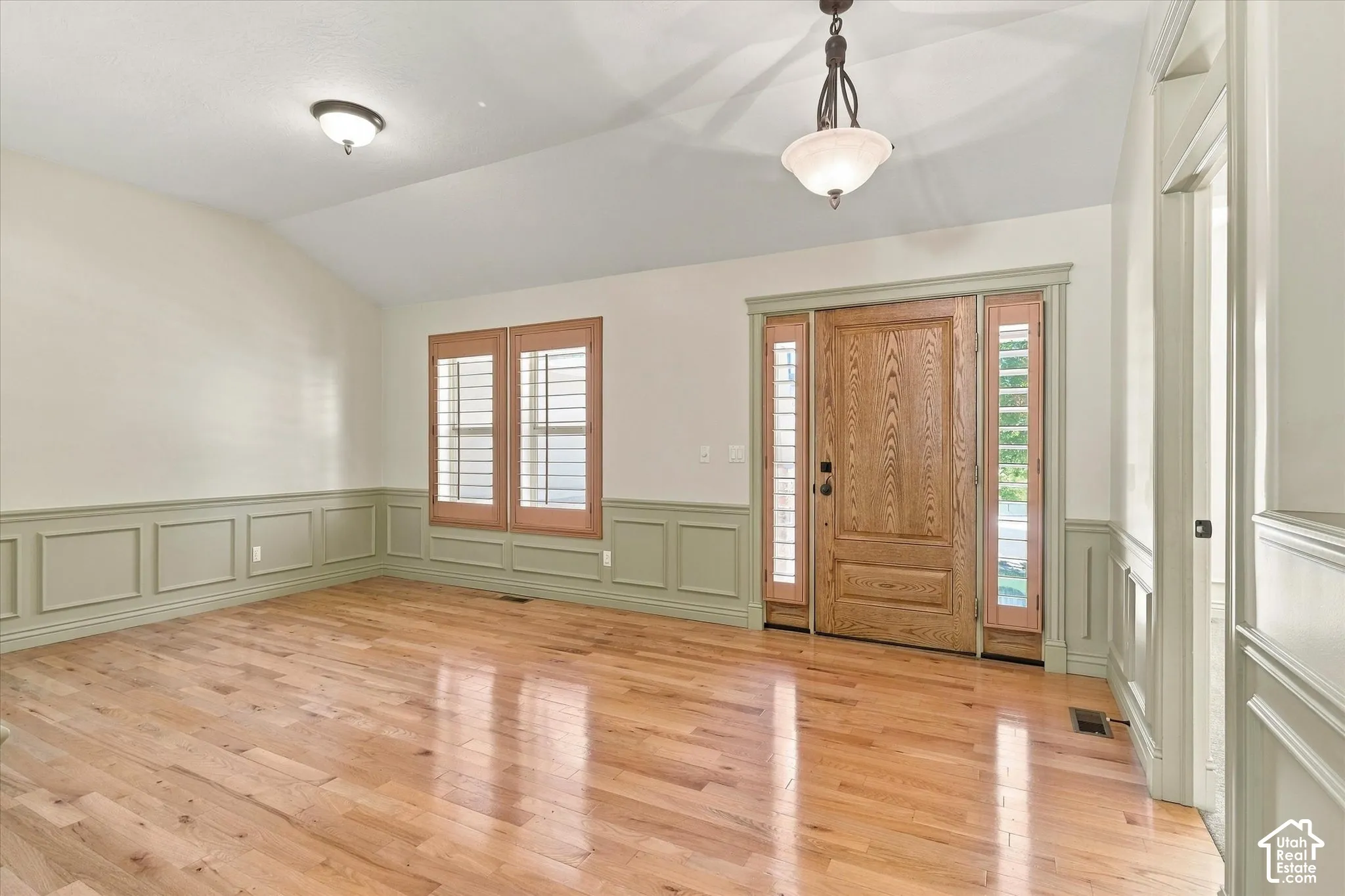 Vaulted ceiling with an attached light fixture, wood-finish flooring, wainscoting, wooden entry door with sidelights, and wooden window shutters