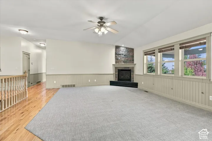 Living area with vaulted ceilings, a ceiling fan, a stone-clad fireplace with a mantel, multiple windows, and partial wood-finish flooring