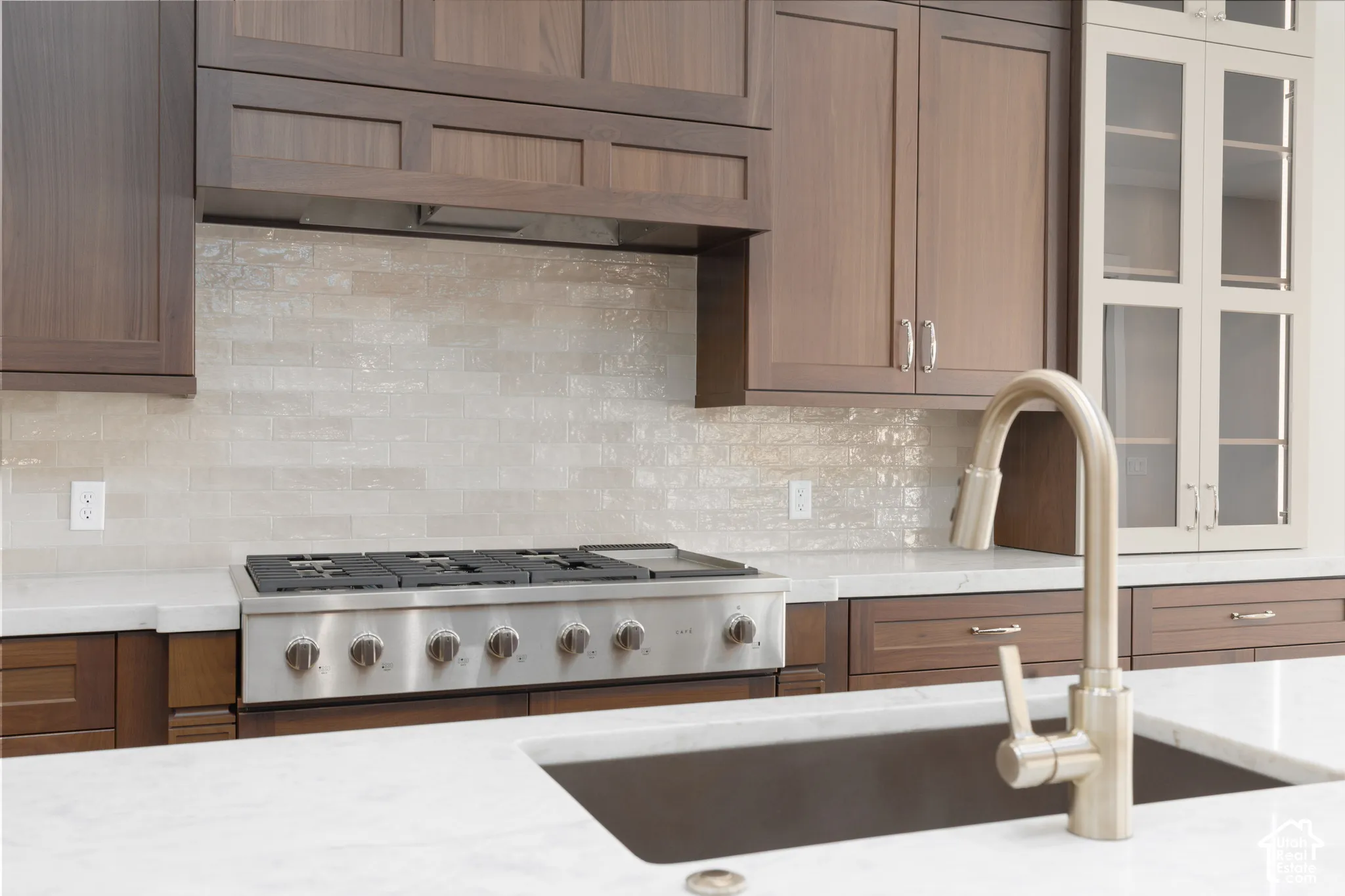 Stainless steel gas range with a tile backsplash, complemented by a large basin sink with a brushed nickel faucet, and wood-finish cabinetry
