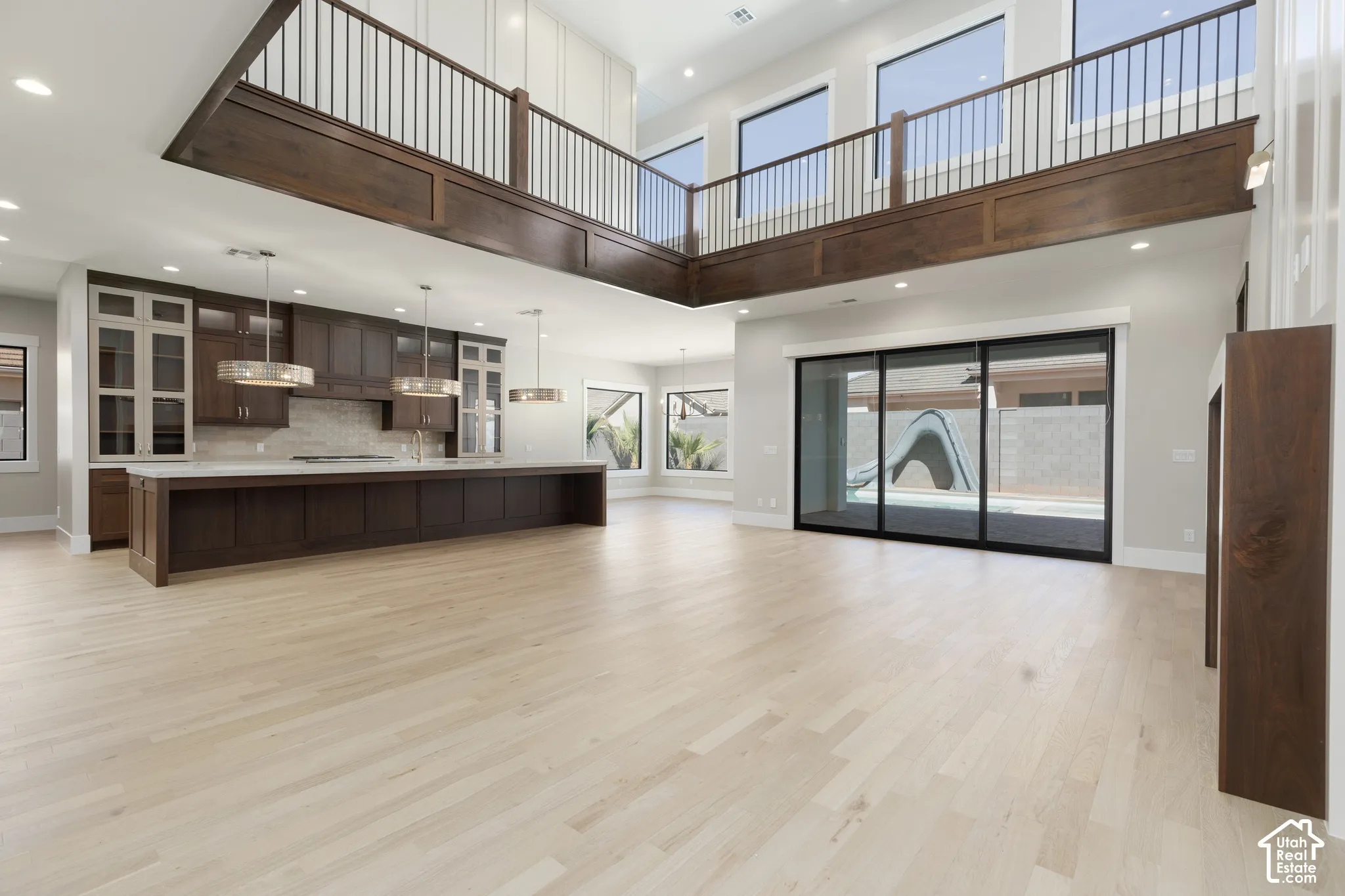 Open-concept living area with light wood-finish flooring, a full kitchen featuring dark wood cabinetry and a large island, and a second-story interior balcony with dark wood and iron railings
