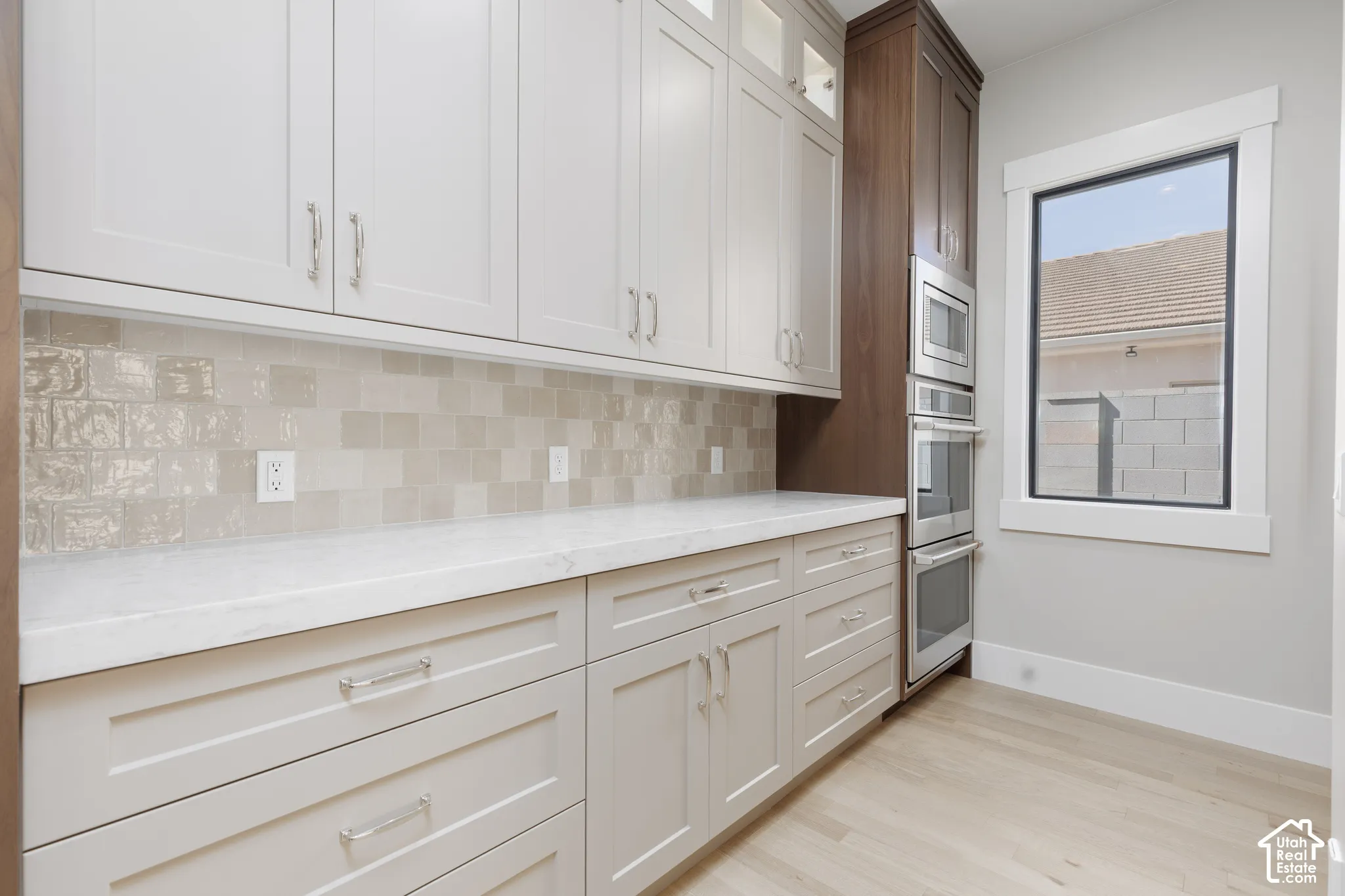 Light-toned cabinetry with coordinating hardware, crisp white countertops, textured tile backsplash, built-in stainless steel appliances, and light wood-finish flooring