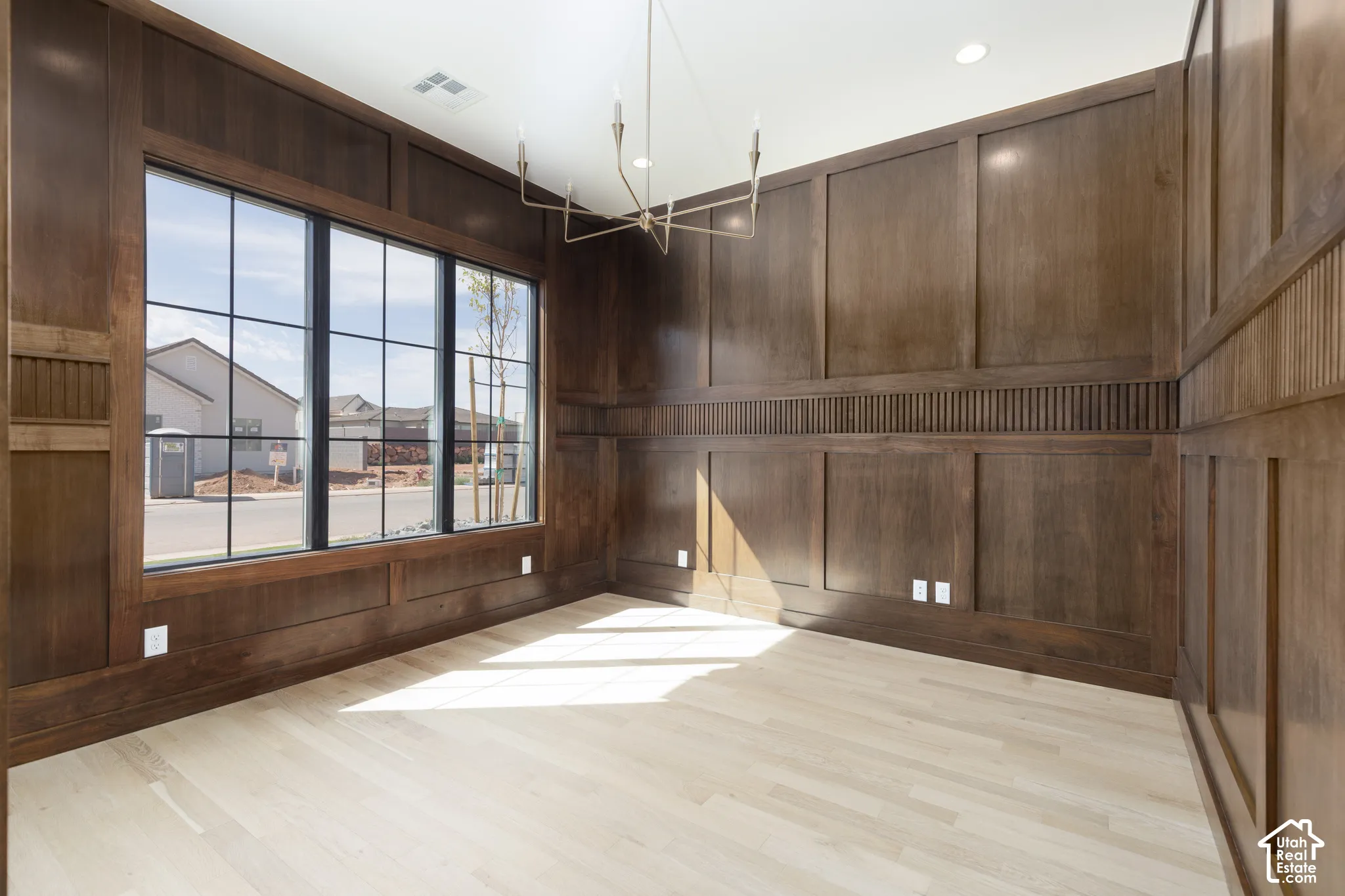 Light wood-finish flooring, dark wood wall paneling, large grid-pane windows, and a modern linear chandelier