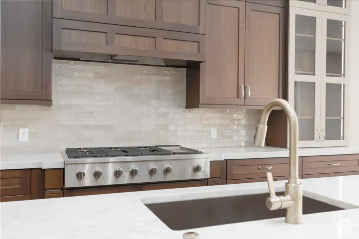 Stainless steel gas range with a tile backsplash, complemented by a large basin sink with a brushed nickel faucet, and wood-finish cabinetry