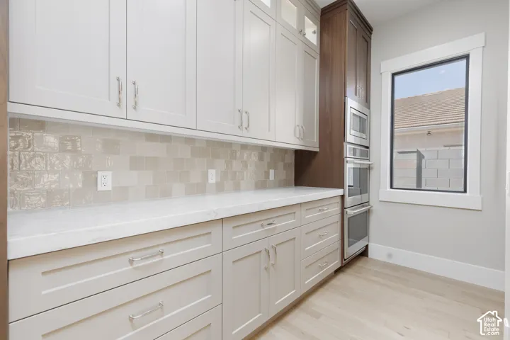 Light-toned cabinetry with coordinating hardware, crisp white countertops, textured tile backsplash, built-in stainless steel appliances, and light wood-finish flooring