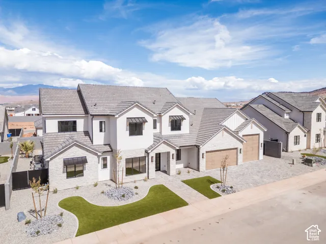 White stucco exterior with light gray brick accents, dark gray tile roof, two-car garage with wood-finish doors, black window awnings, and a paver driveway