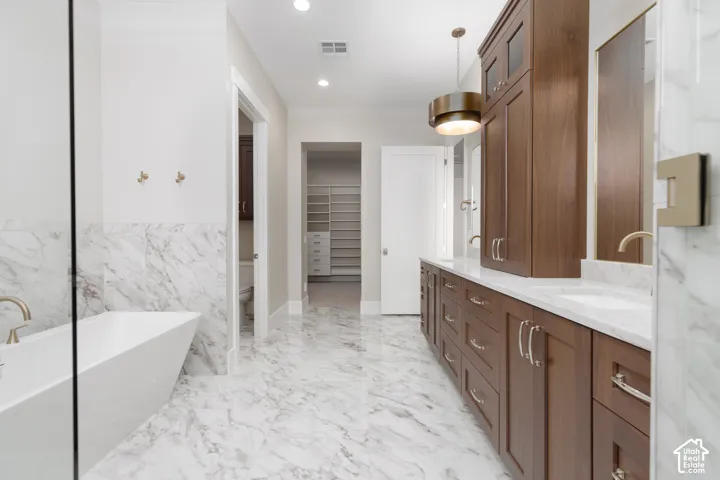 Freestanding soaking tub, marble-patterned floor and wall tile, dual vanity with undermount sinks, wood-finish cabinetry, and recessed lighting