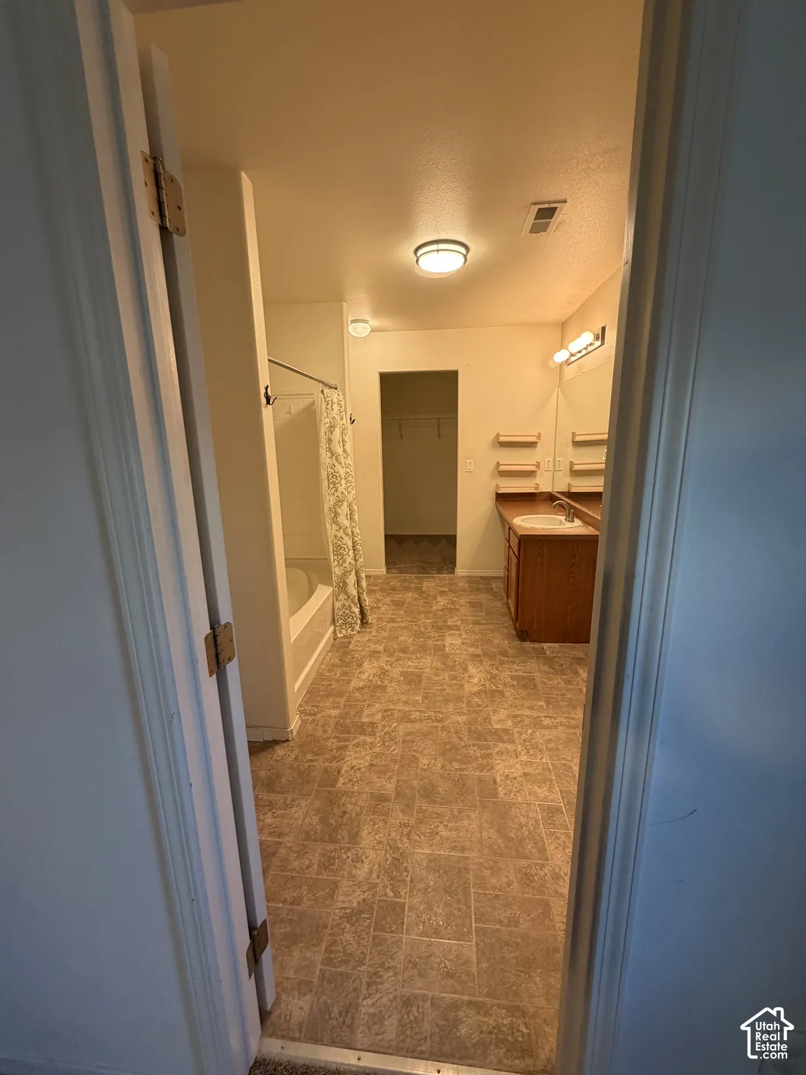 Brown patterned tile flooring, built-in wooden vanity with single basin sink, wall-mounted mirror with integrated lighting, and a white bathtub with shower