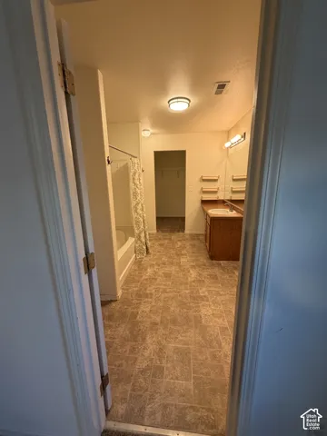 Brown patterned tile flooring, built-in wooden vanity with single basin sink, wall-mounted mirror with integrated lighting, and a white bathtub with shower
