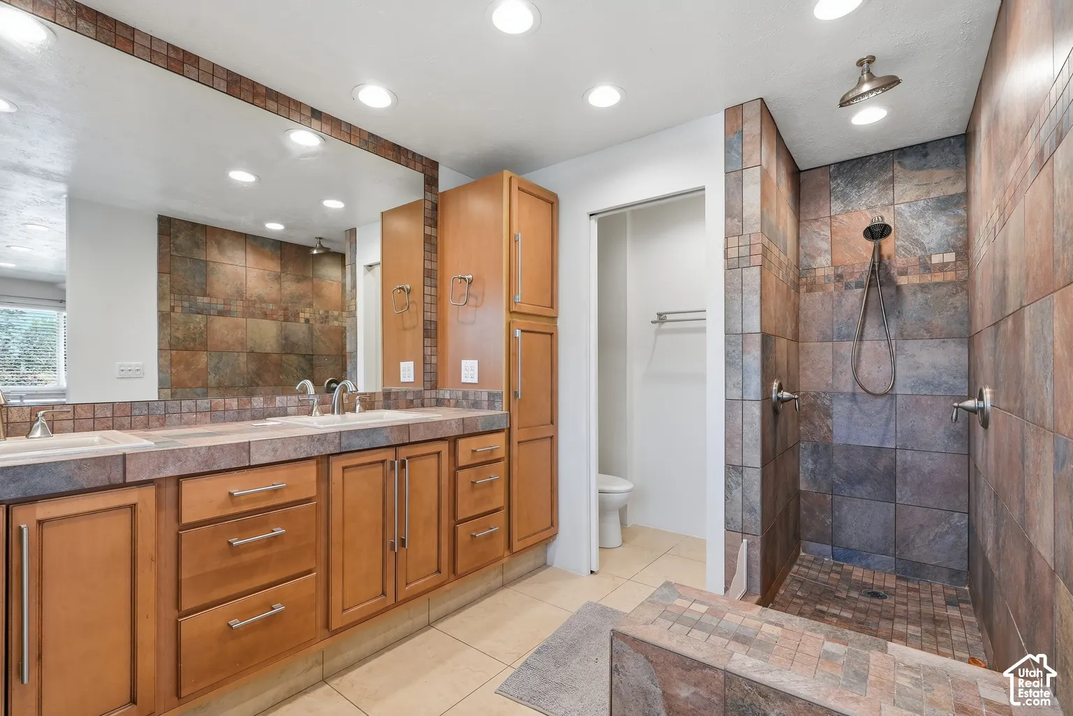 Dual vanity with integrated sinks, wood-finish cabinetry, a full-width mirror, a tiled walk-in shower with dual shower heads, and recessed lighting