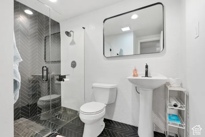 Walk-in shower with clear glass enclosure and dark herringbone tile, white pedestal sink with matte black faucet, and a rectangular black-framed mirror