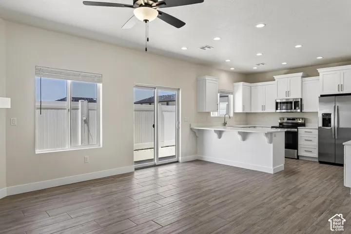 Open-concept living space featuring wood-finish flooring, a contemporary kitchen with white cabinetry, stainless steel appliances, a breakfast bar, and a ceiling fan