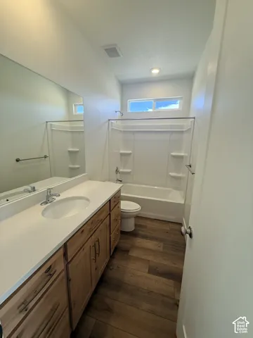 Full-width mirror, wood-finish flooring, white vanity countertop, light wood-tone cabinetry, and a bathtub with shower