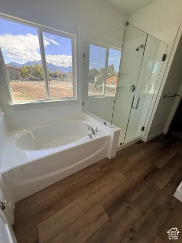 Soaking tub with deck-mounted fixtures, glass-enclosed shower, wood-finish flooring, and large windows