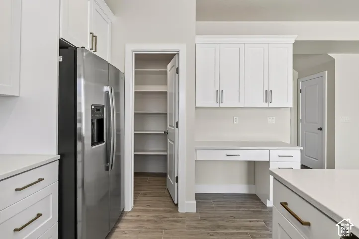 White cabinetry with gold-tone hardware, built-in stainless steel refrigerator, dedicated pantry with shelving, wood-finish flooring, and a built-in desk area with countertop